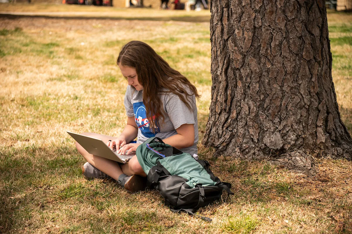A student works on a laptop under a tree