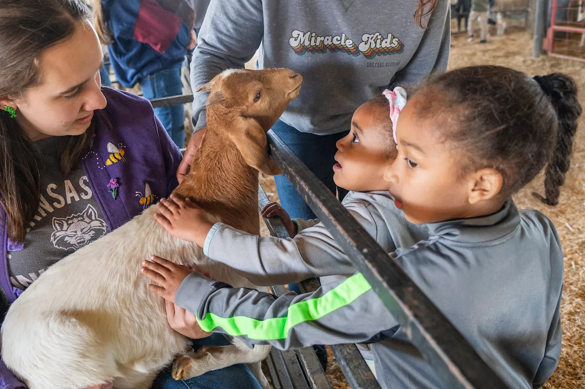 Two young children pet a goat while guided by adults during a farm visit.