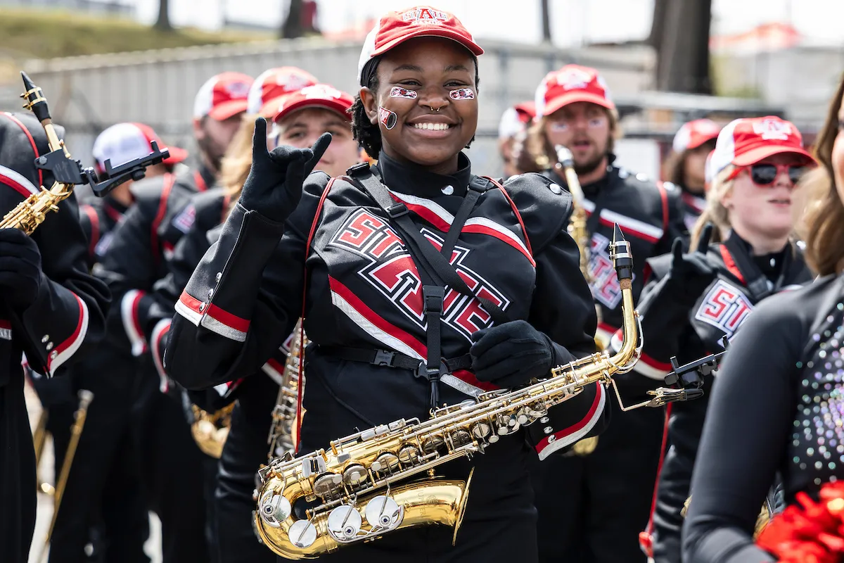 A-State marching band saxophonist smiles and flashes a hand sign while marching before a game.