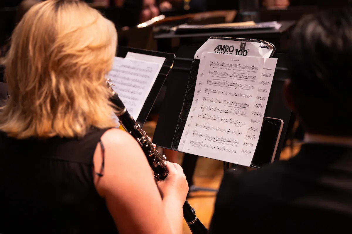 Concert band members seated on stage following a performance moment inside a formal recital hall.