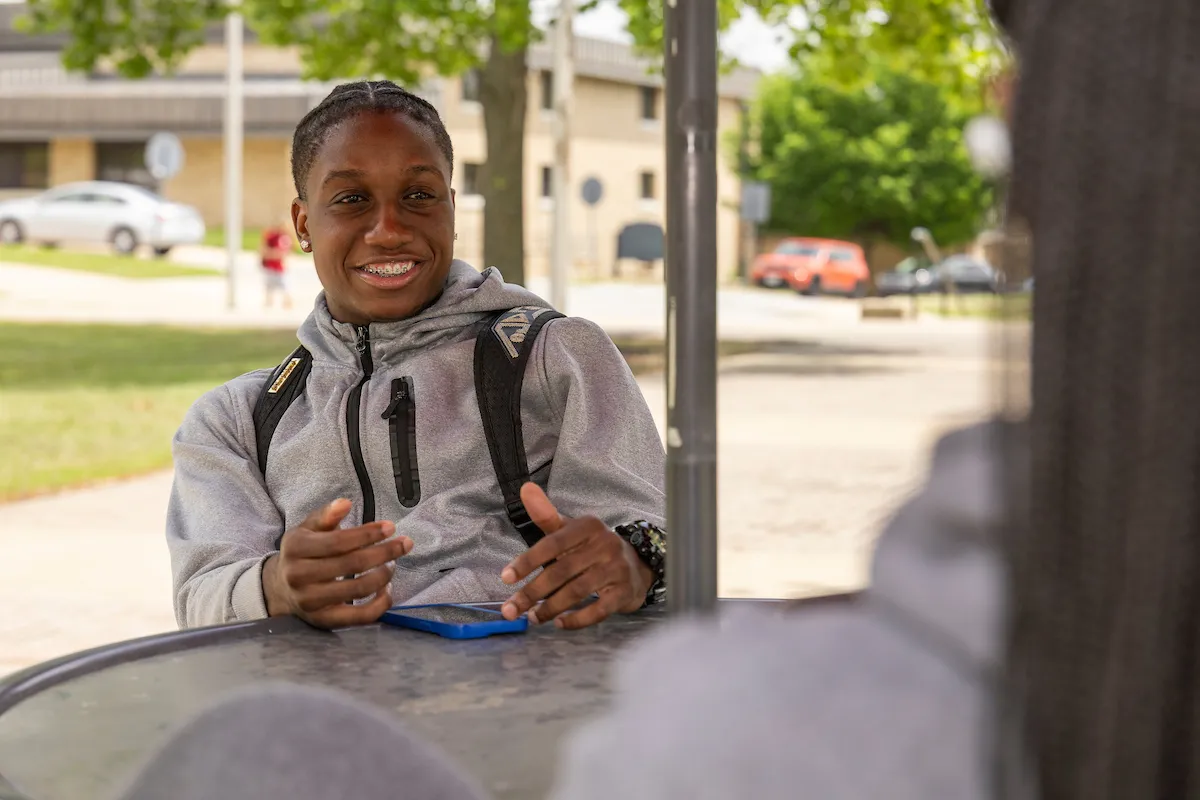 A student visits with another student at an outdoor table.