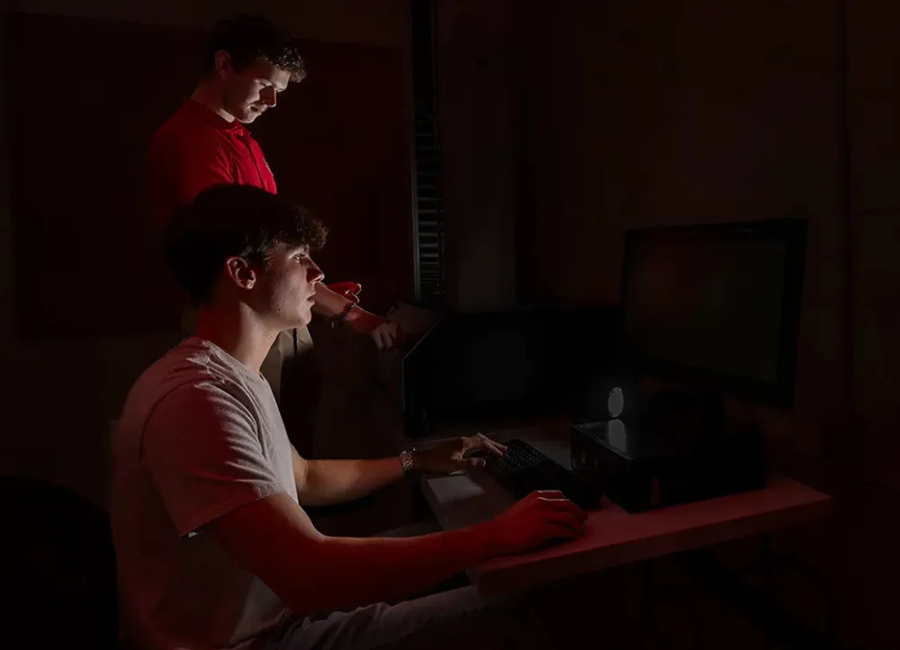 AState students working in a dim lit computer lab with one student looking over the shoulder of the other.
