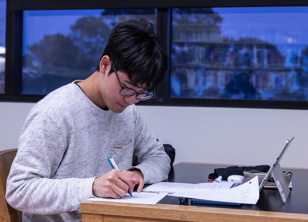 An A-State student focuses on notes while studying with a laptop and papers at a table inside a campus building.