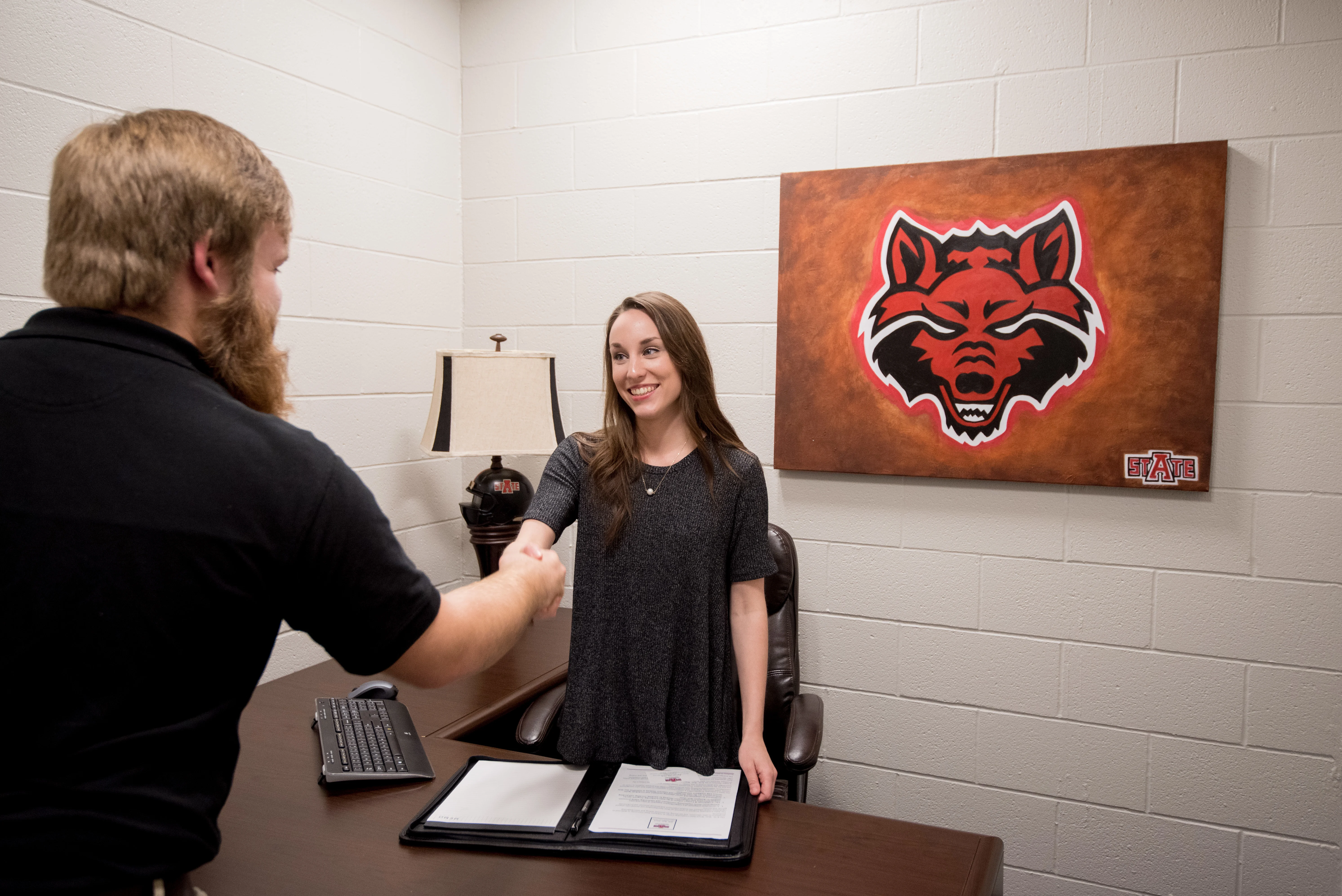 A-State students doing a mock interview in a business class in a staged office with large redwolf poster on the wall.