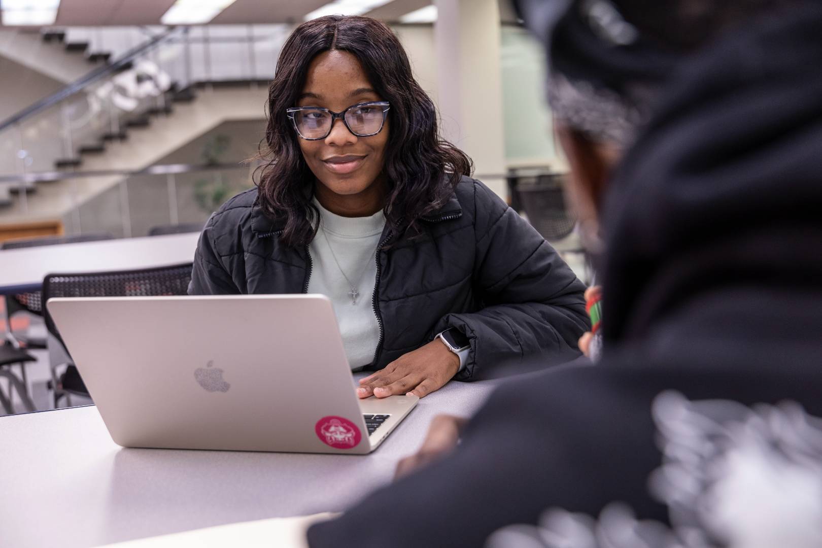 A student sitting at a desk with her laptop and smiling at someone in the foreground