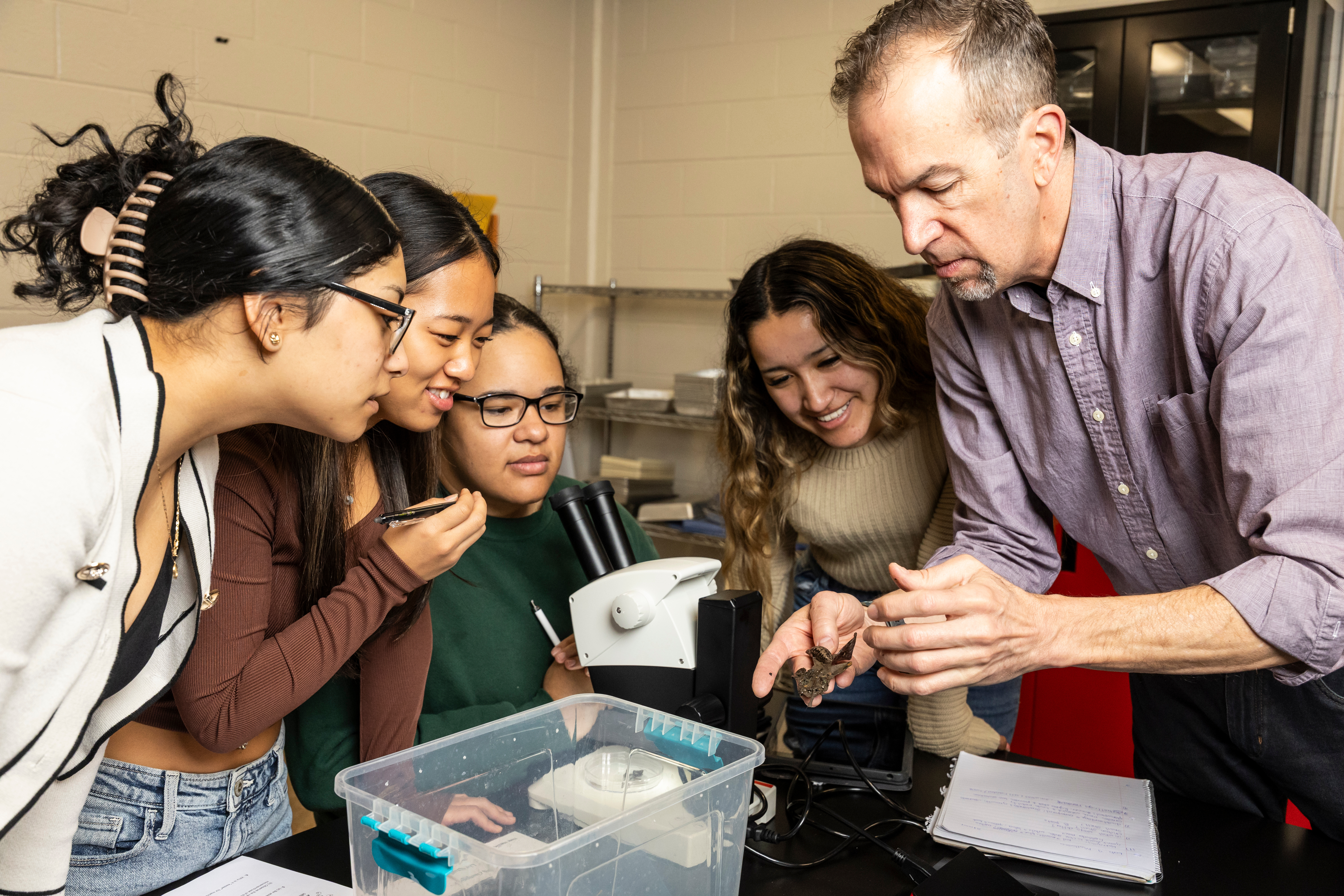 Smiling students huddled around a professor as he explains a specimen to them.