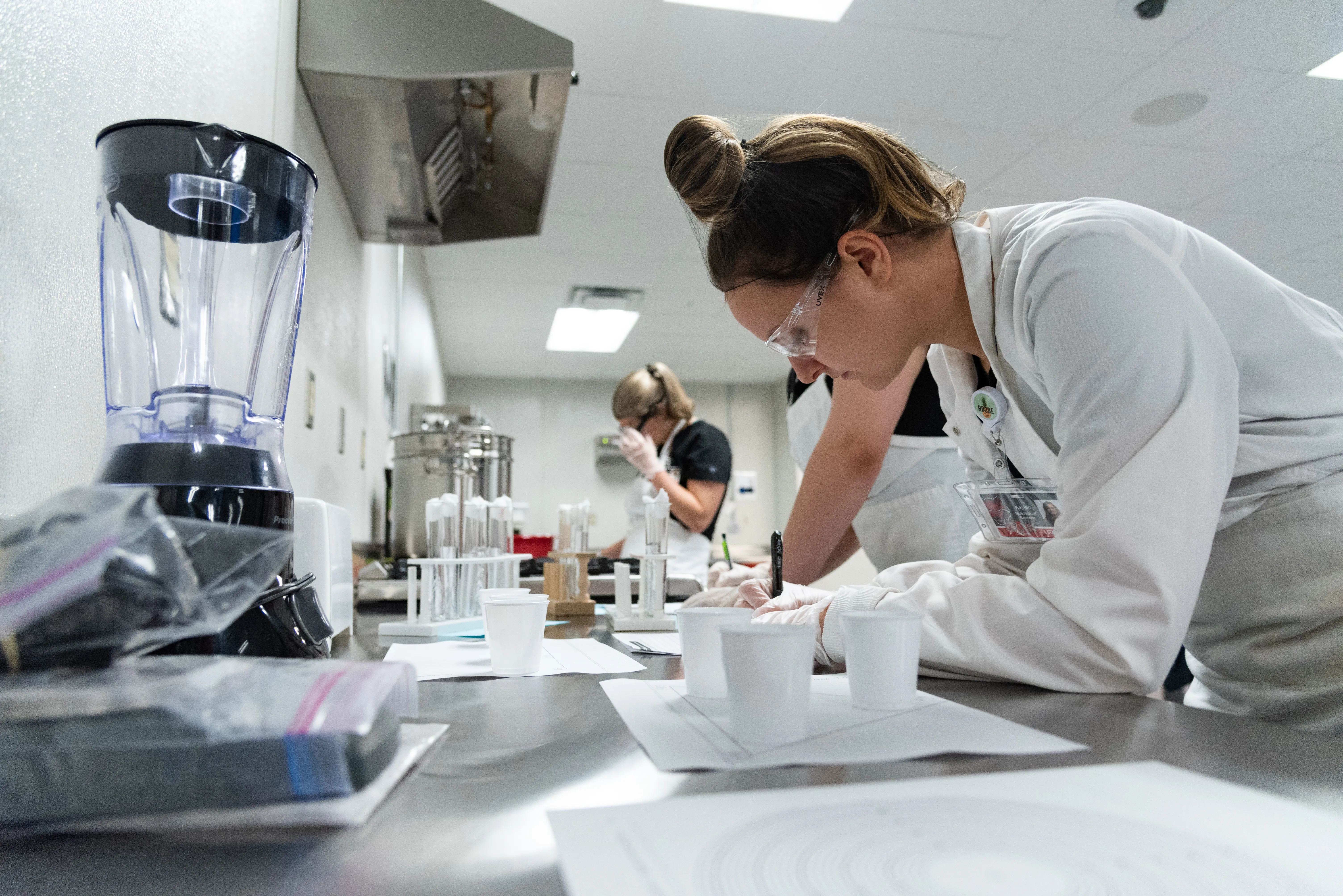 Nutrition student measuring out liquids in a lab coat with beakers and a blender around.