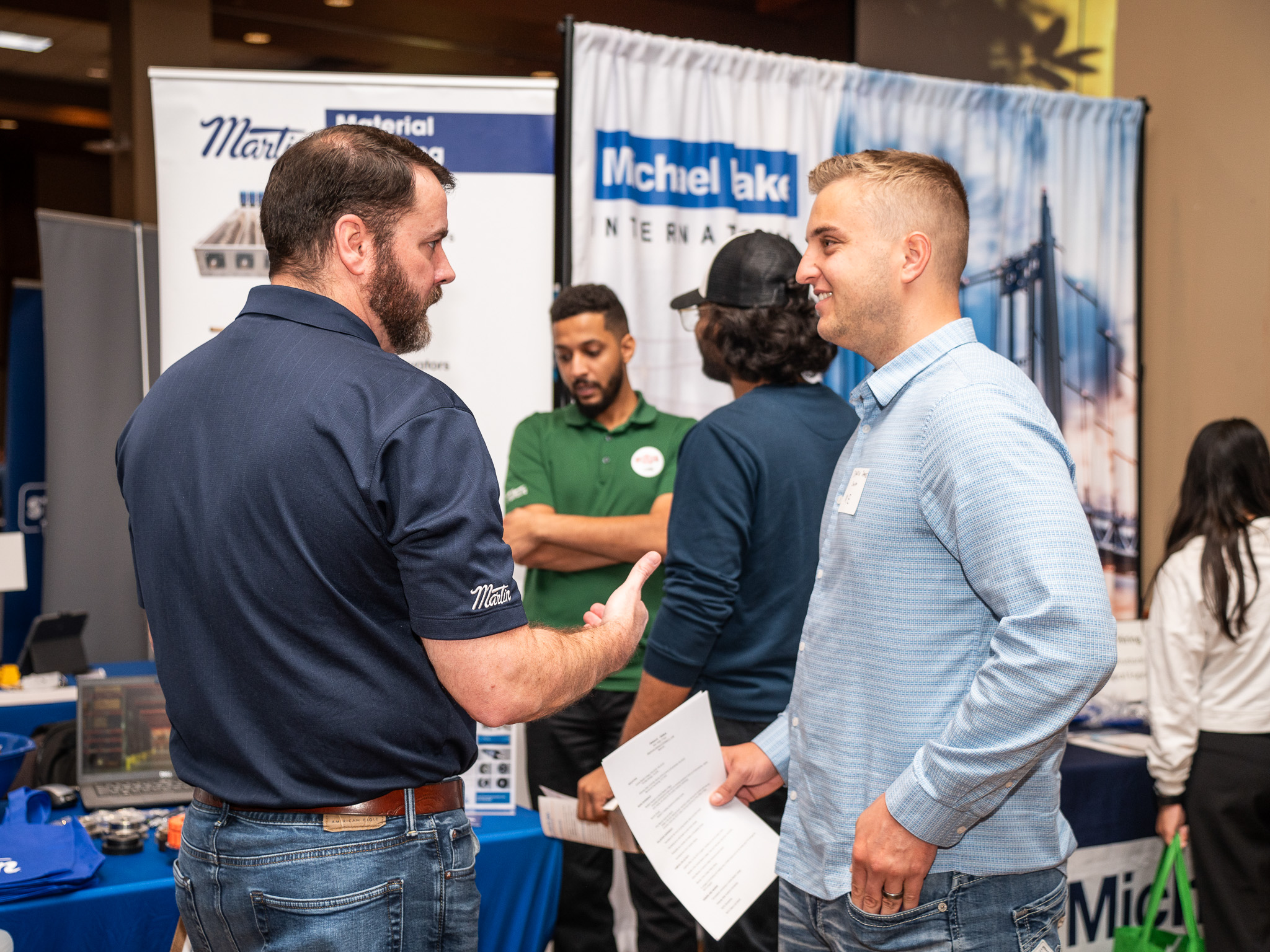 “Several people talking at an indoor networking or employer fair, with two individuals in the foreground holding papers and conversing in front of exhibitor booths with company banners.”