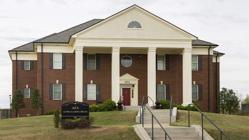Two-story red brick sorority house with white columns and a cream-colored pediment. A black sign in front reads “ΑΓΔ Alpha Gamma Delta.” Steps lead up to a red front door, and the house is surrounded by grass and shrubs.