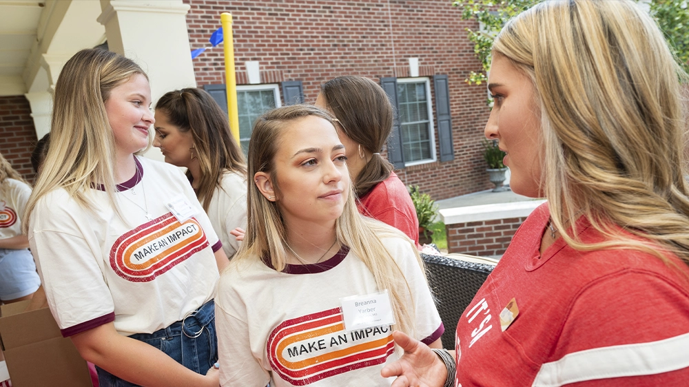 Group of individuals wearing “Make an Impact” shirts talking together outside a brick building during recruitment.