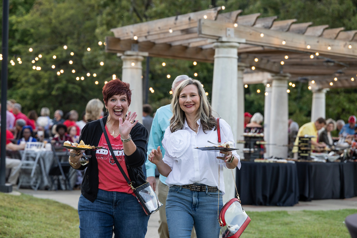 Two women smile and carry food at an evening outdoor Arkansas State alumni celebration.