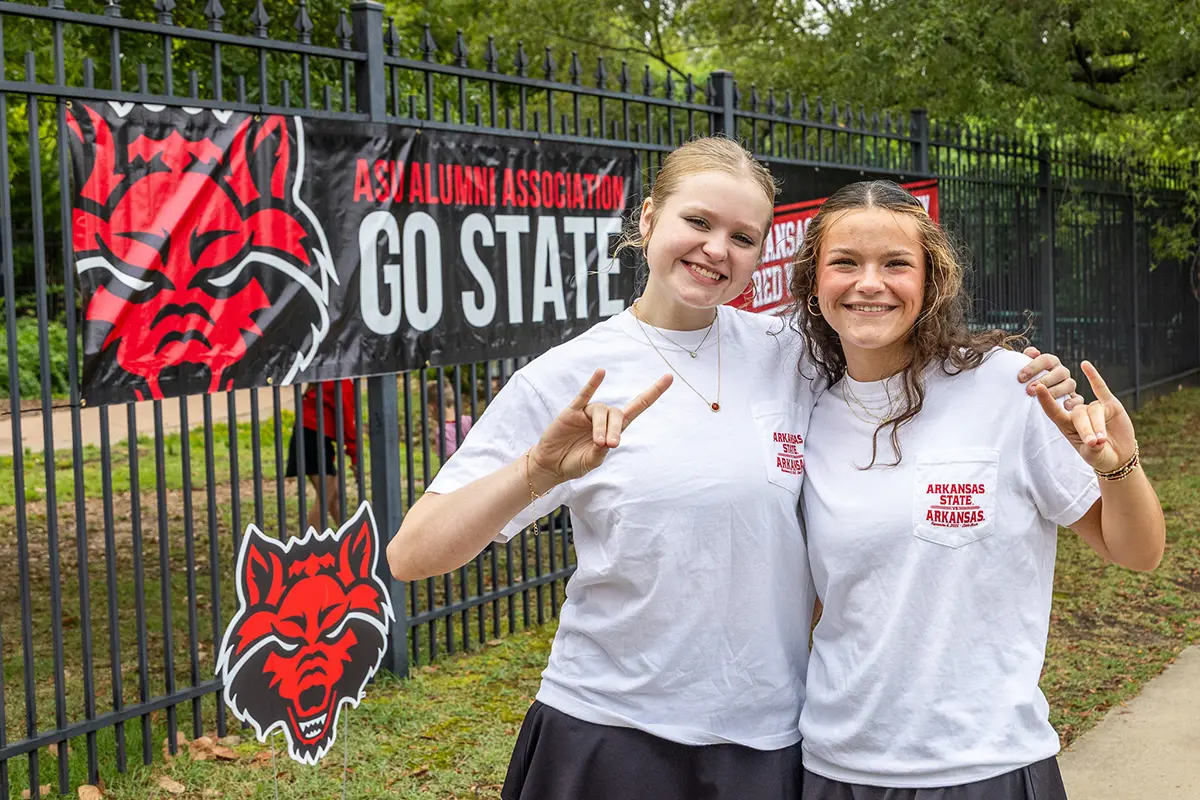 Two Arkansas State University students smile and pose in front of a campus fence decorated with a Red Wolves banner that reads “ASU Alumni Association – Go State.” Both wear white A-State shirts and make the Wolves Up hand sign, showing school spirit.