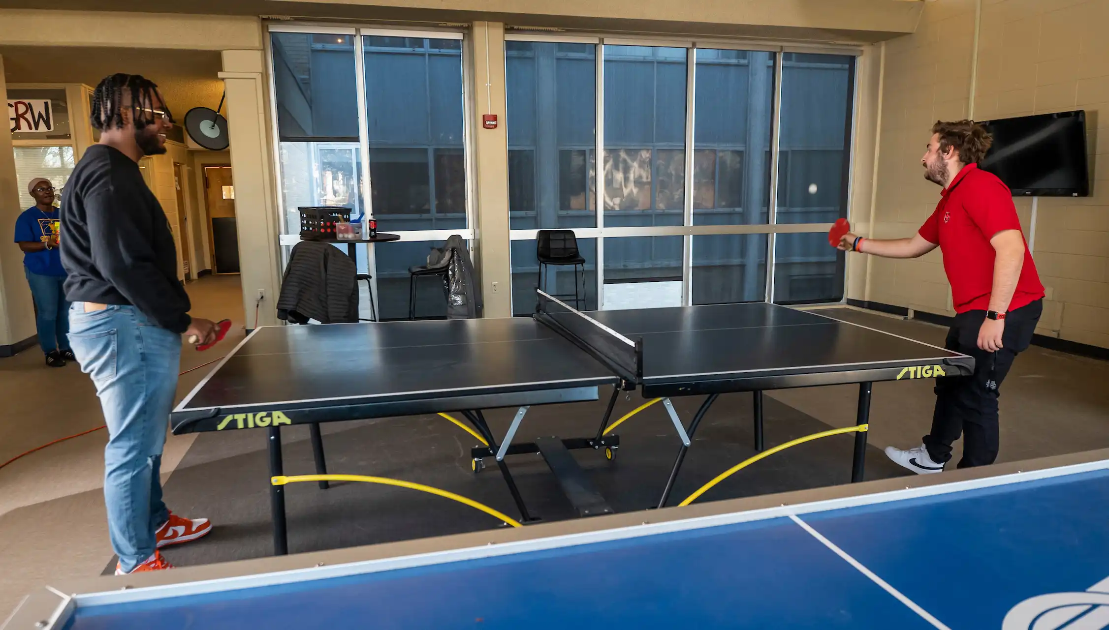 Two students playing ping pong in a lobby with a wall of windows in the background.