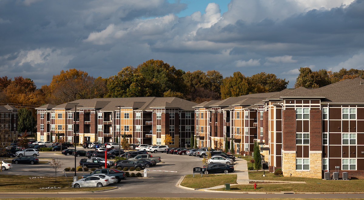 Modern A-State student apartments with fall trees and full parking lot under cloudy sky.