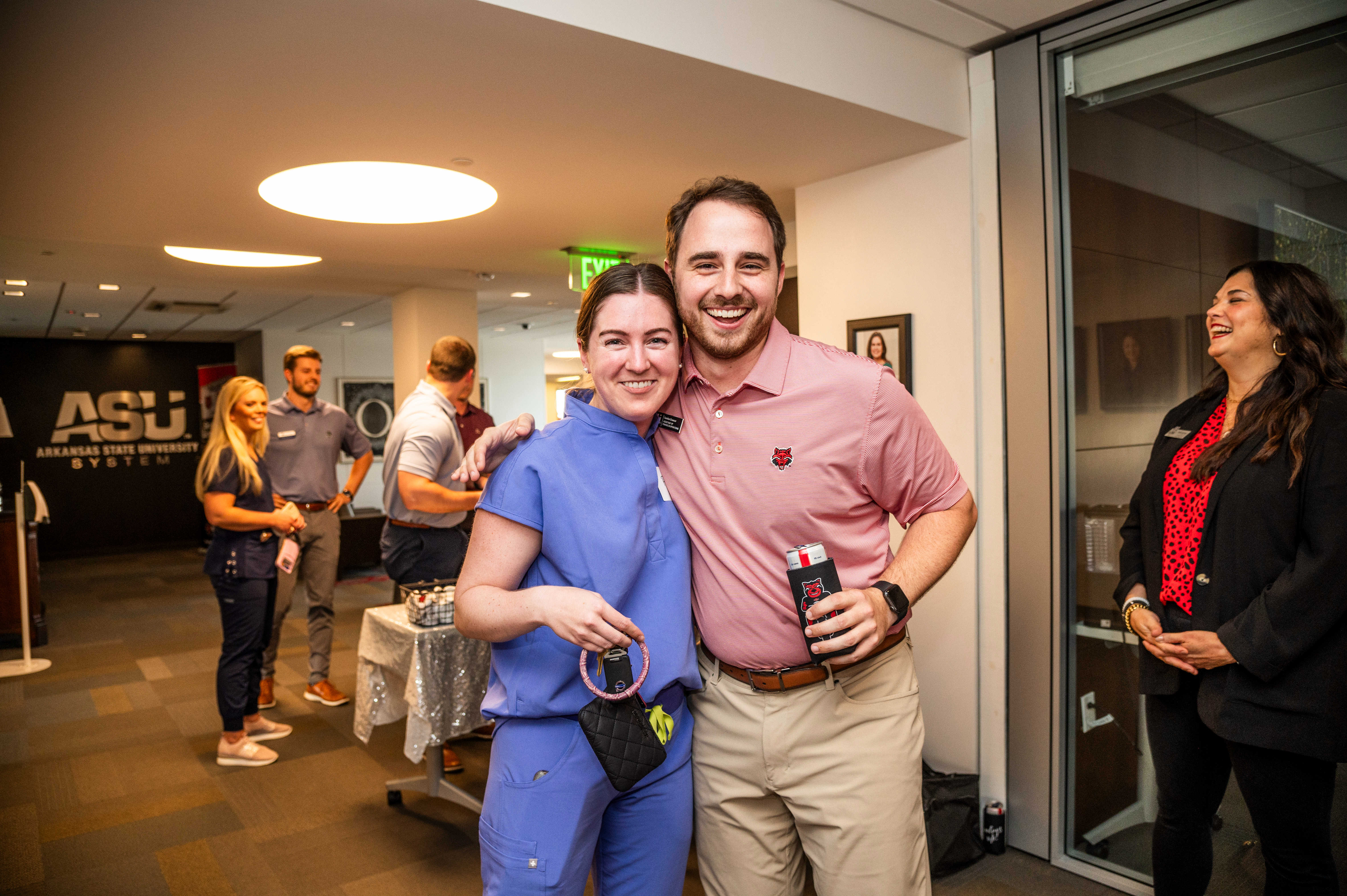 Two A-State alumni pose together at a social event with others mingling in the background at the ASU System Office.