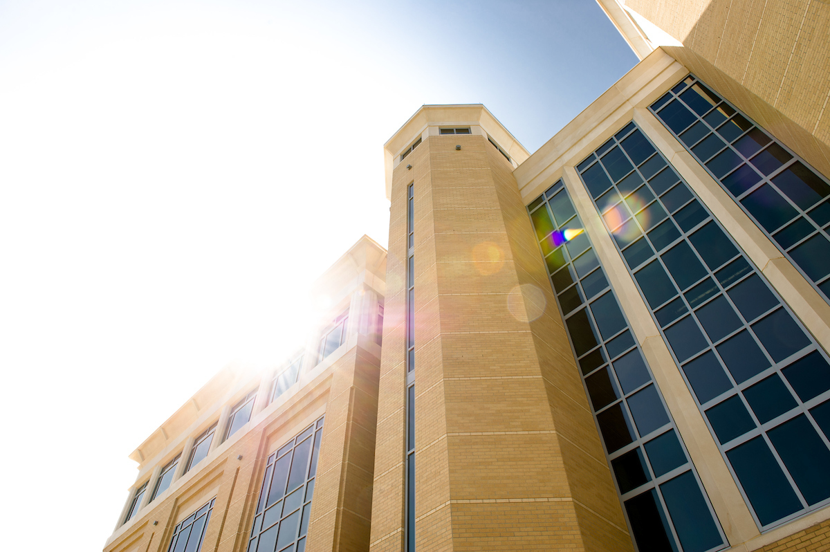 Sunlight flares over a tall A-State campus building with large windows and tan brick exterior.