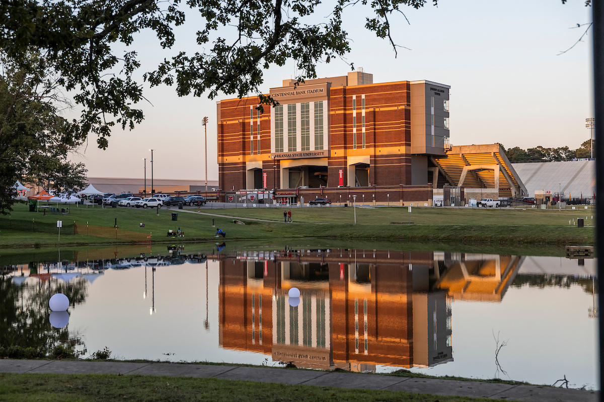Centennial Bank Stadium is reflected in the pond at sunset with tailgaters and tents in the background.