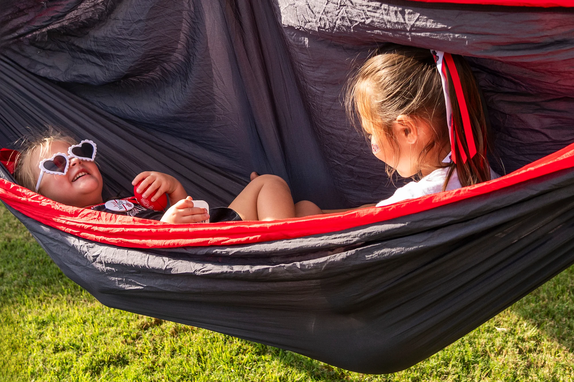 Two children smiling and playing in a scarlet and black hammock. 