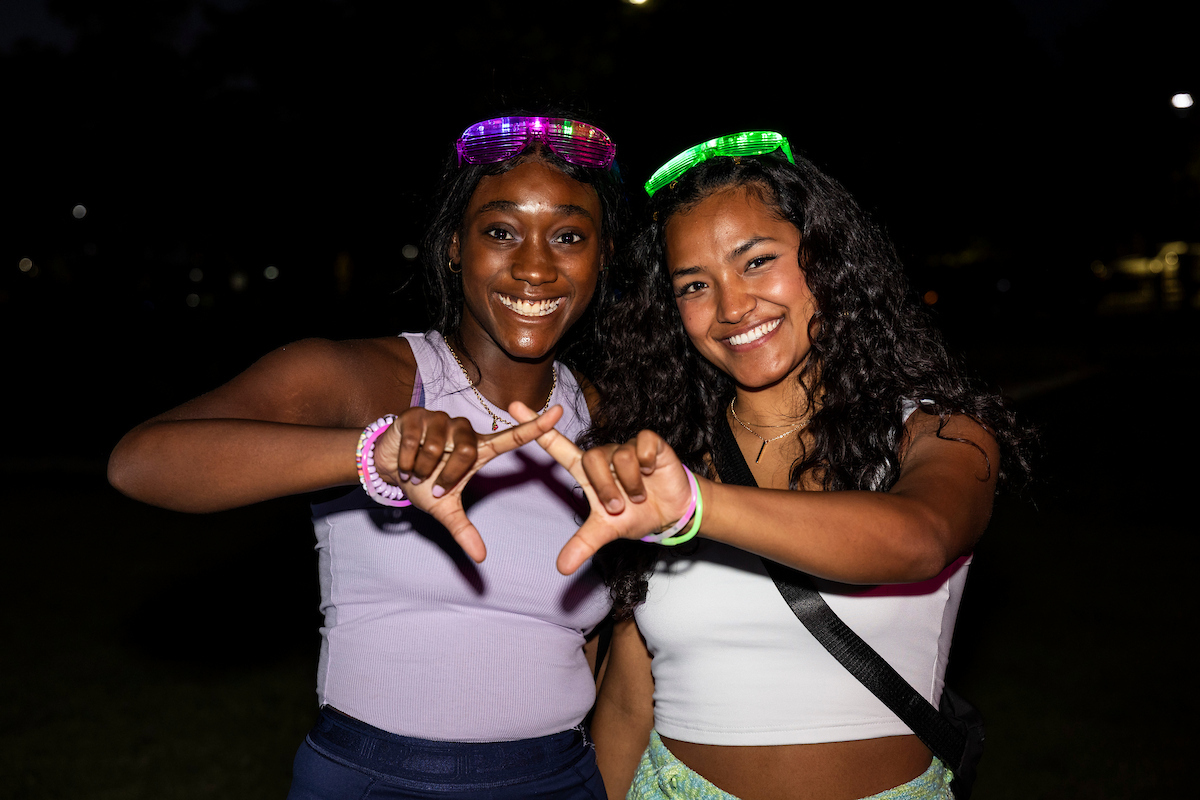 Two A-State students smile under glow lights at night, wearing neon glasses during a campus glow event.