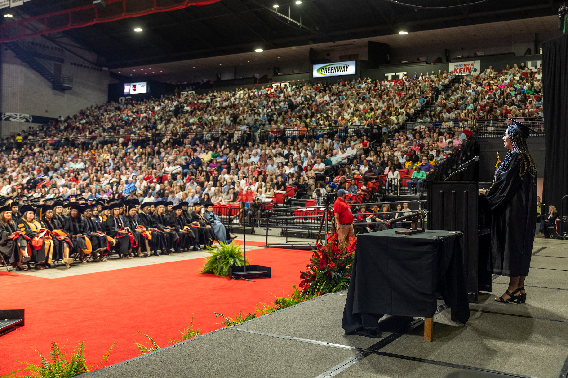 A-State graduate speaks on stage during commencement with a large crowd seated.