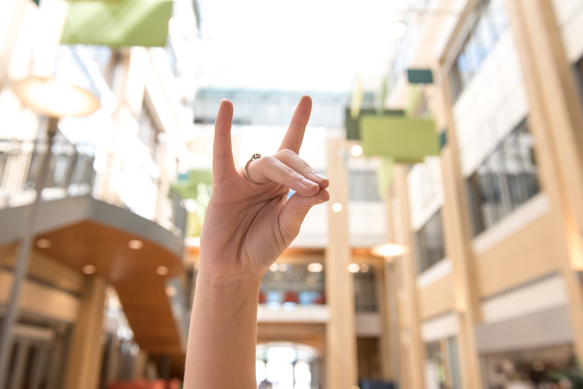 Close-up of a hand showing the Wolves Up sign inside a brightly lit A-State campus building.