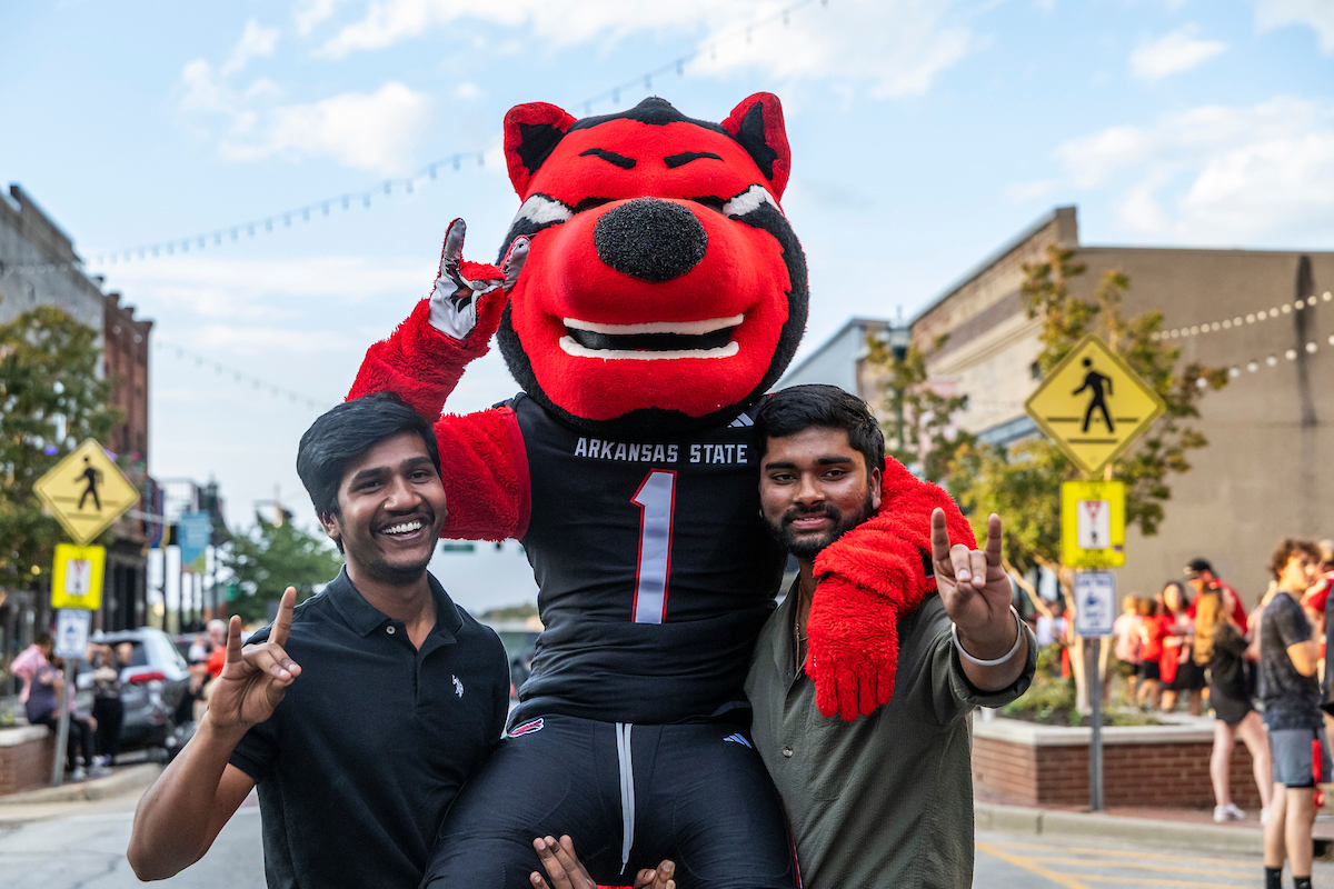 Two A-State students pose with Howl mascot during a spirited downtown event celebrating Red Wolves pride.