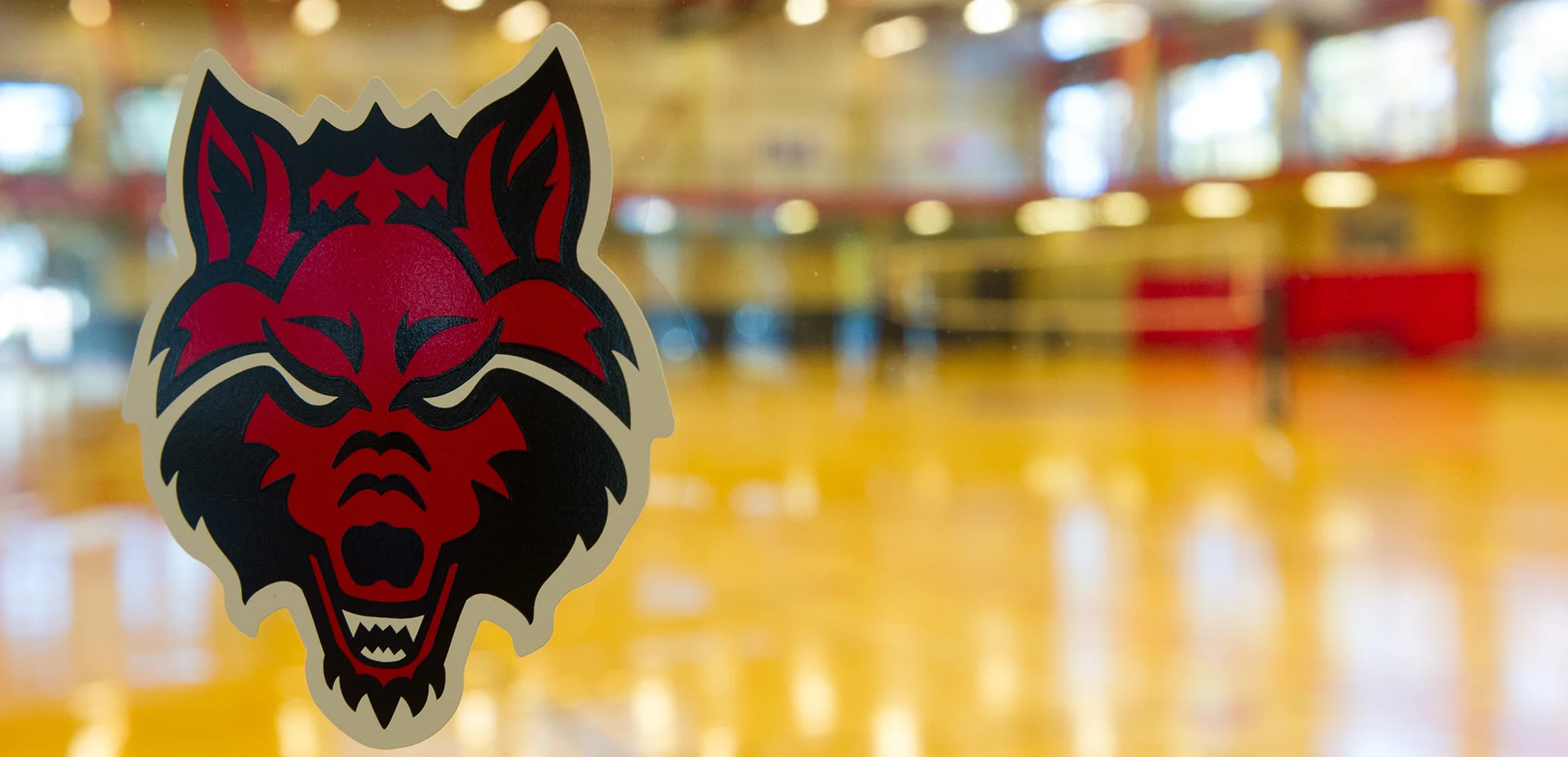 The Red Wolves logo on a pane of glass with a basketball court out of focus in the background.