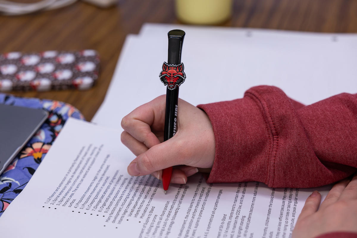 Close-up of a student writing notes with an AState Red Wolves pen during a study session with printed documents and school supplies on the table.