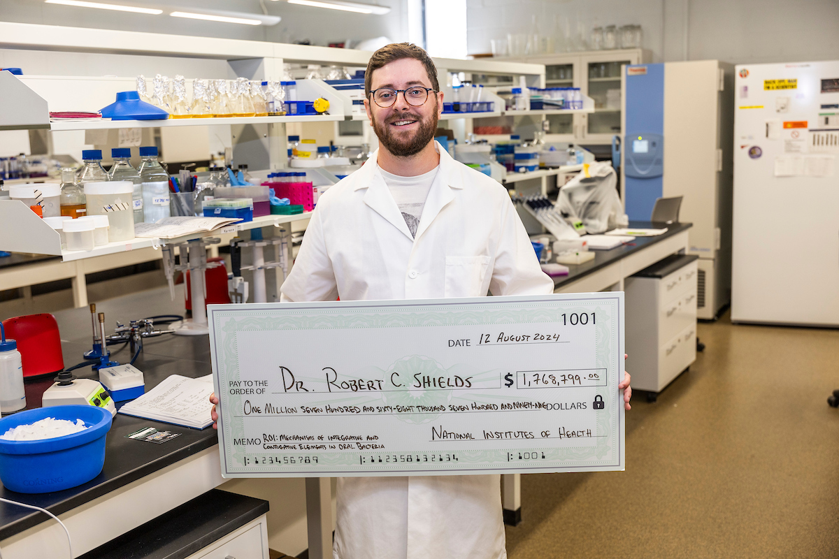 An A-State researcher in a university laboratory holds a giant check to fund his project.