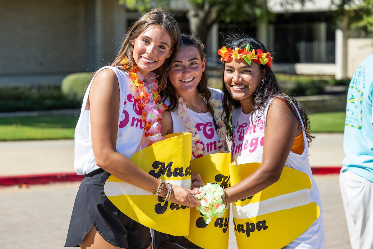 Three A-State students celebrate sorority bid day in Chi Omega shirts with leis and yellow signs, smiling and holding hands outside on campus.