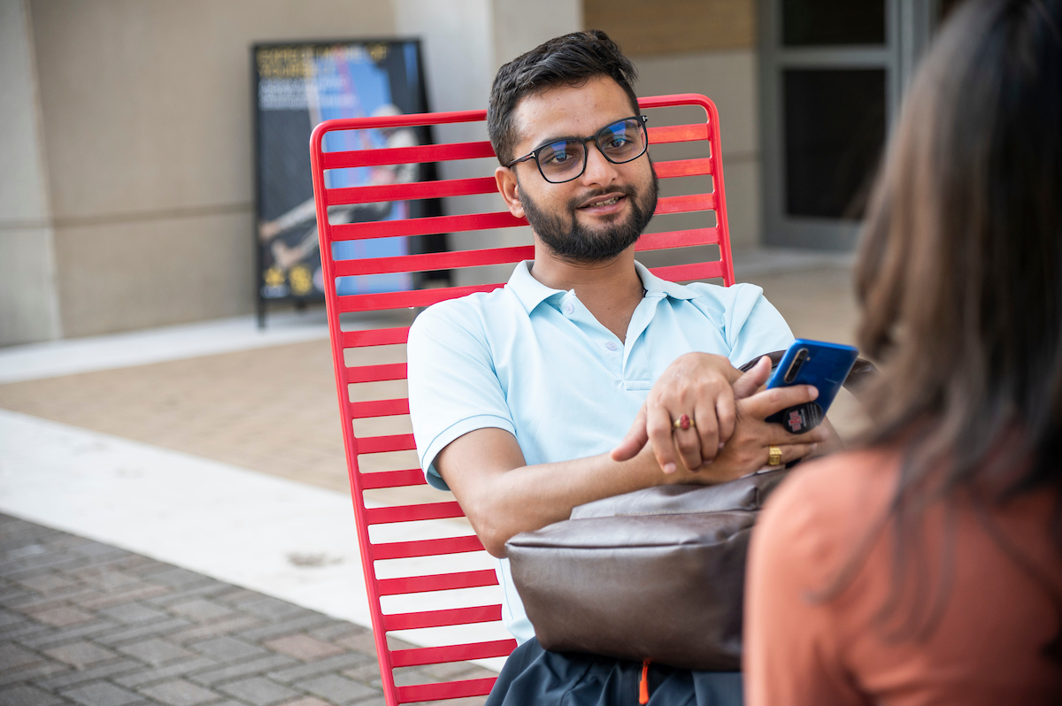 A student relaxes in a red chair and chats with a friend on a campus patio.