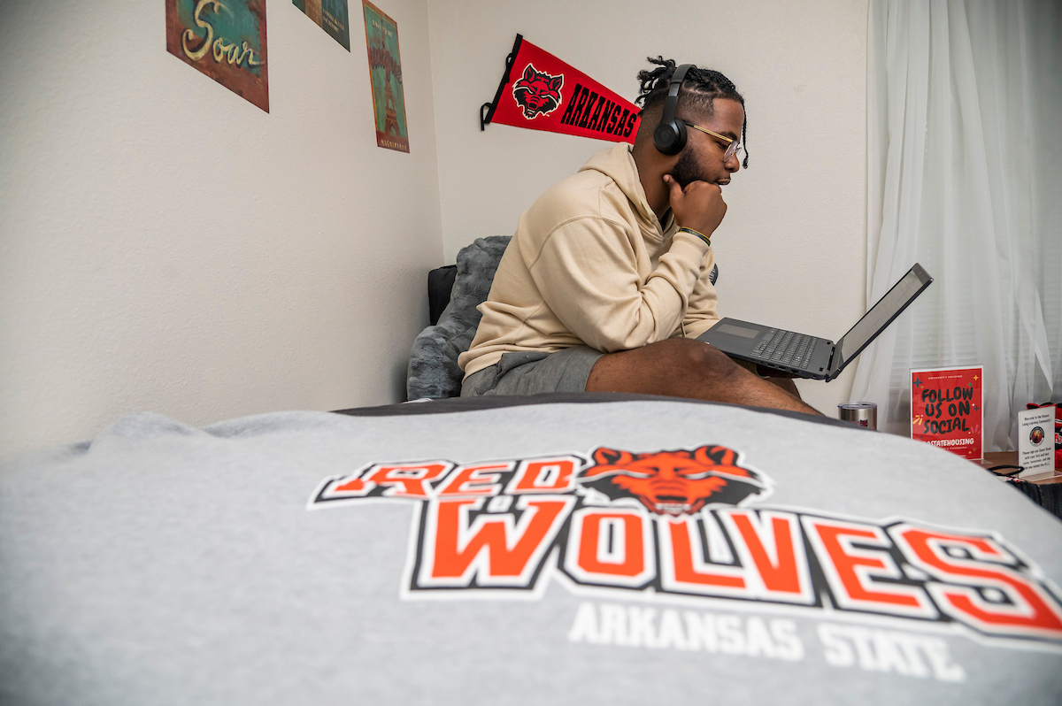A-State student studies online in campus housing with Red Wolves logo and dorm room decor in the background.