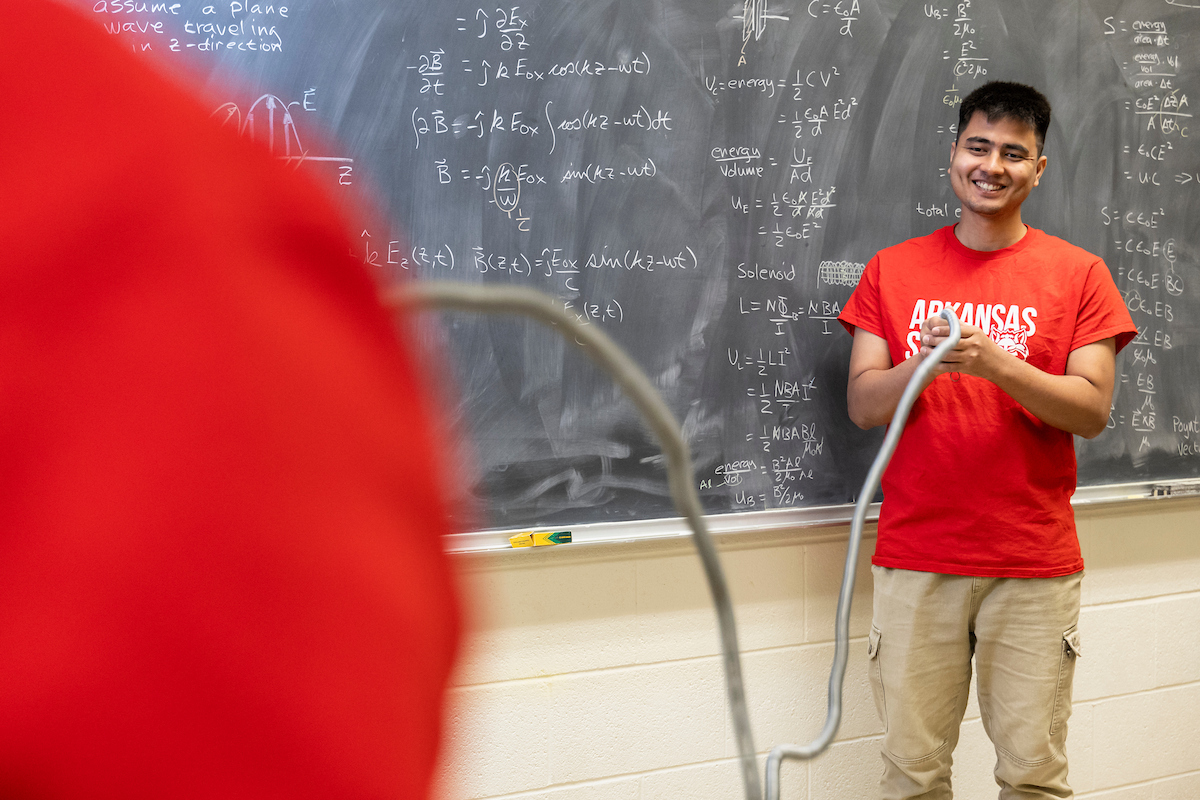 A-State student leads physics demo in front of chalkboard with equations.