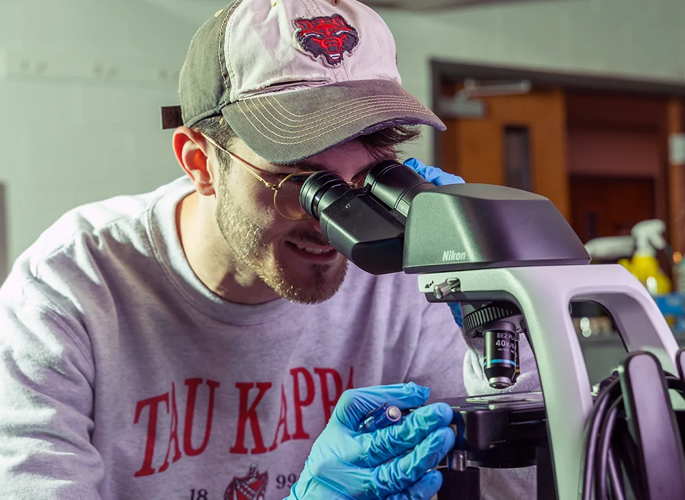 Student in Red Wolves hat using microscope.