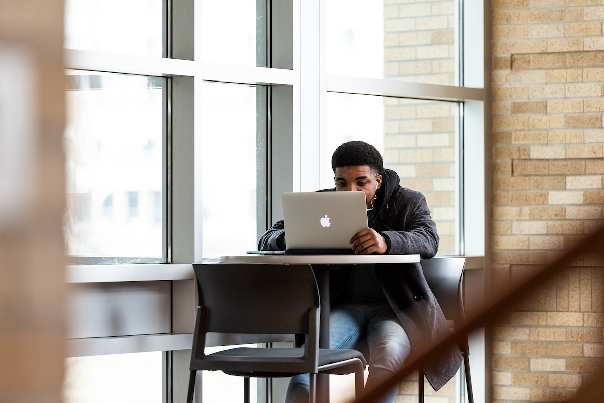 A-State student focused on his laptop while studying by large windows on campus.