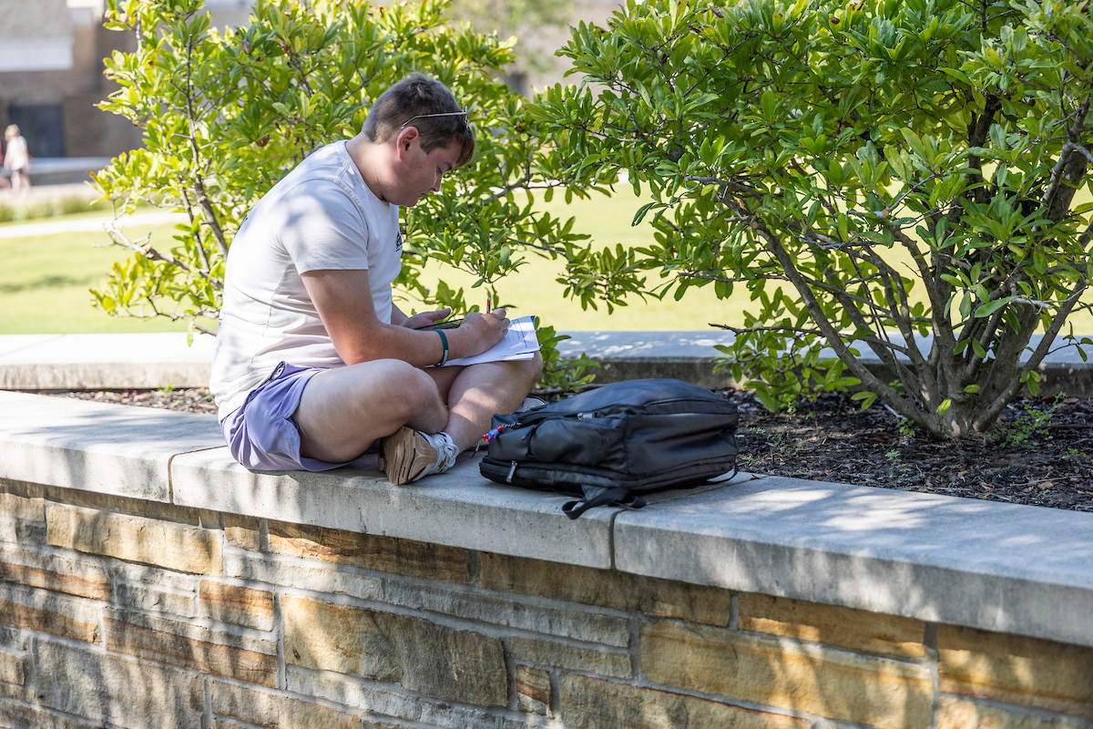 A-State student sitting on a ledge outside taking notes in a notebook.