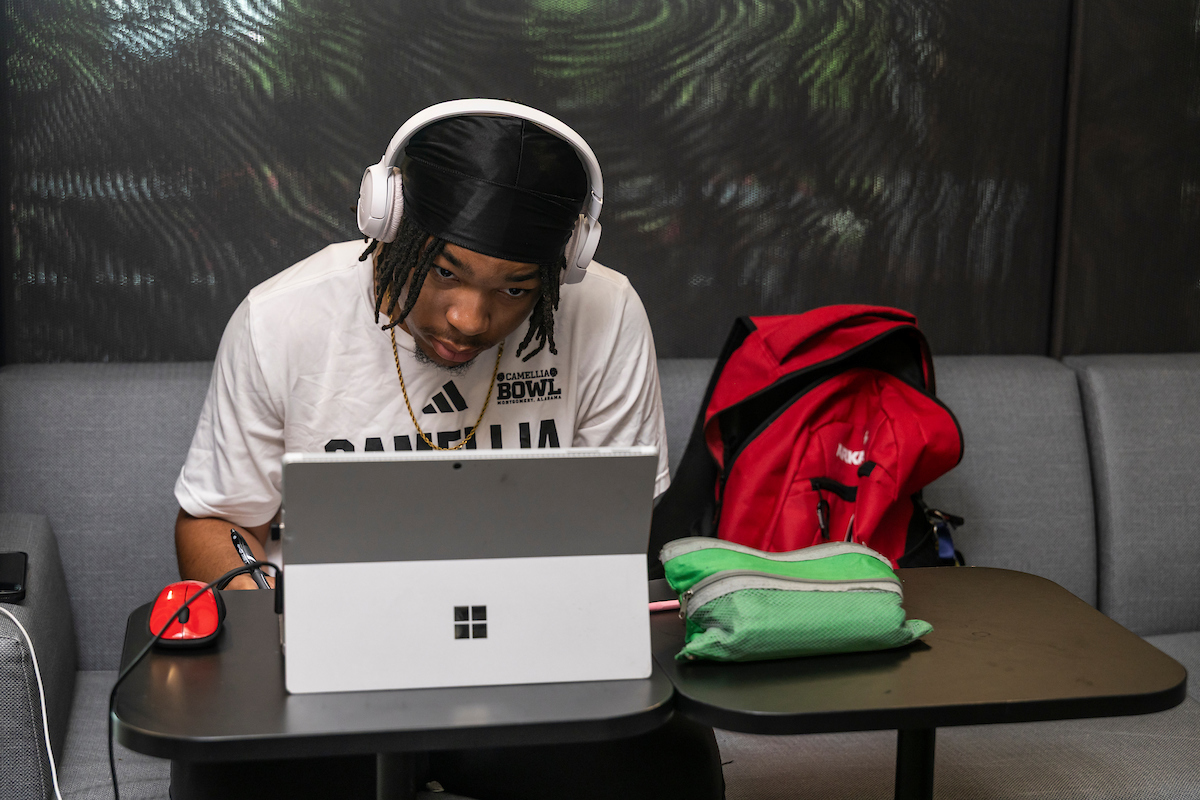 A-State student studies on a tablet while wearing headphones, seated indoors with a red backpack and pencil case nearby.