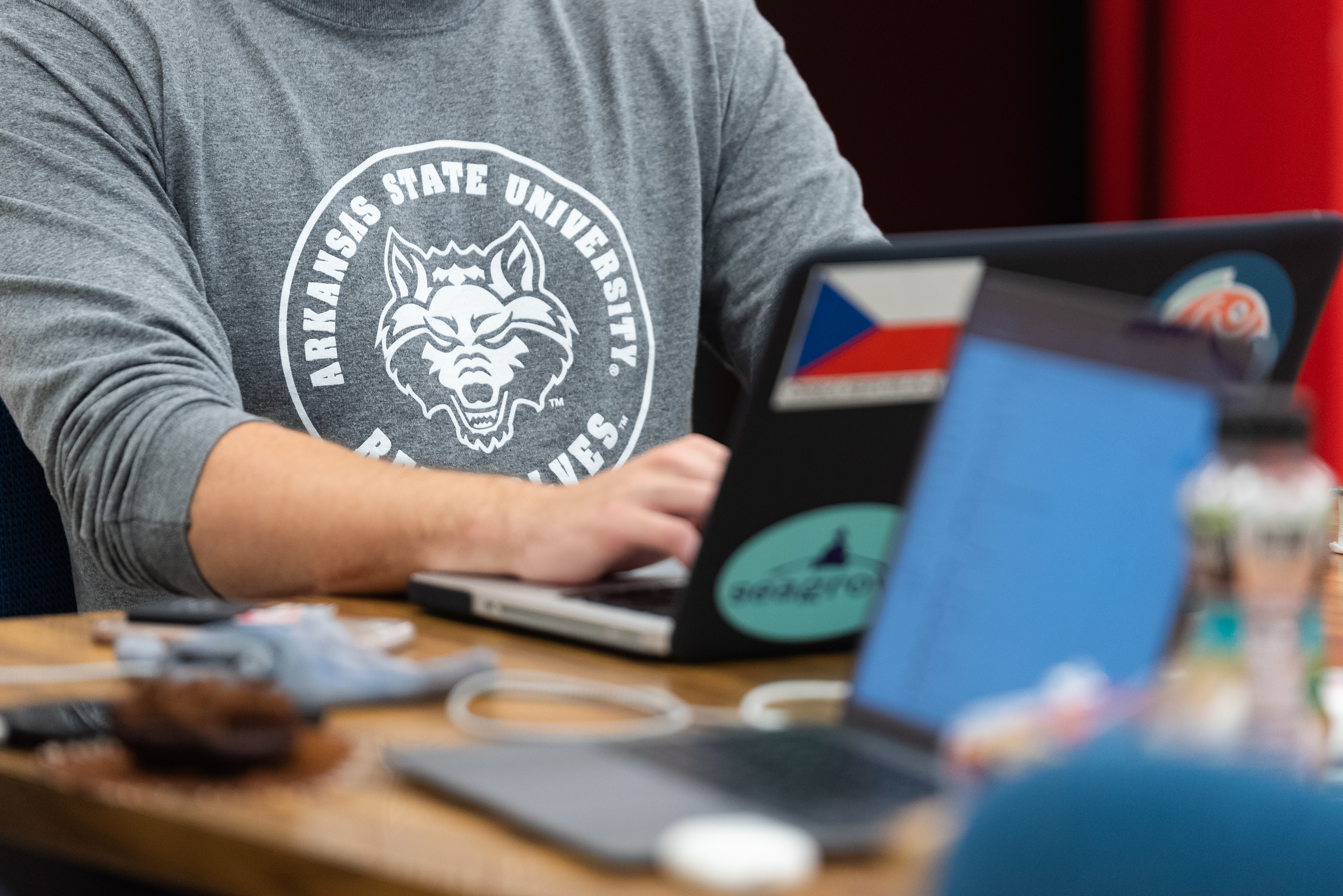 Close-up of student in Red Wolves shirt working on laptop with stickers at a desk.