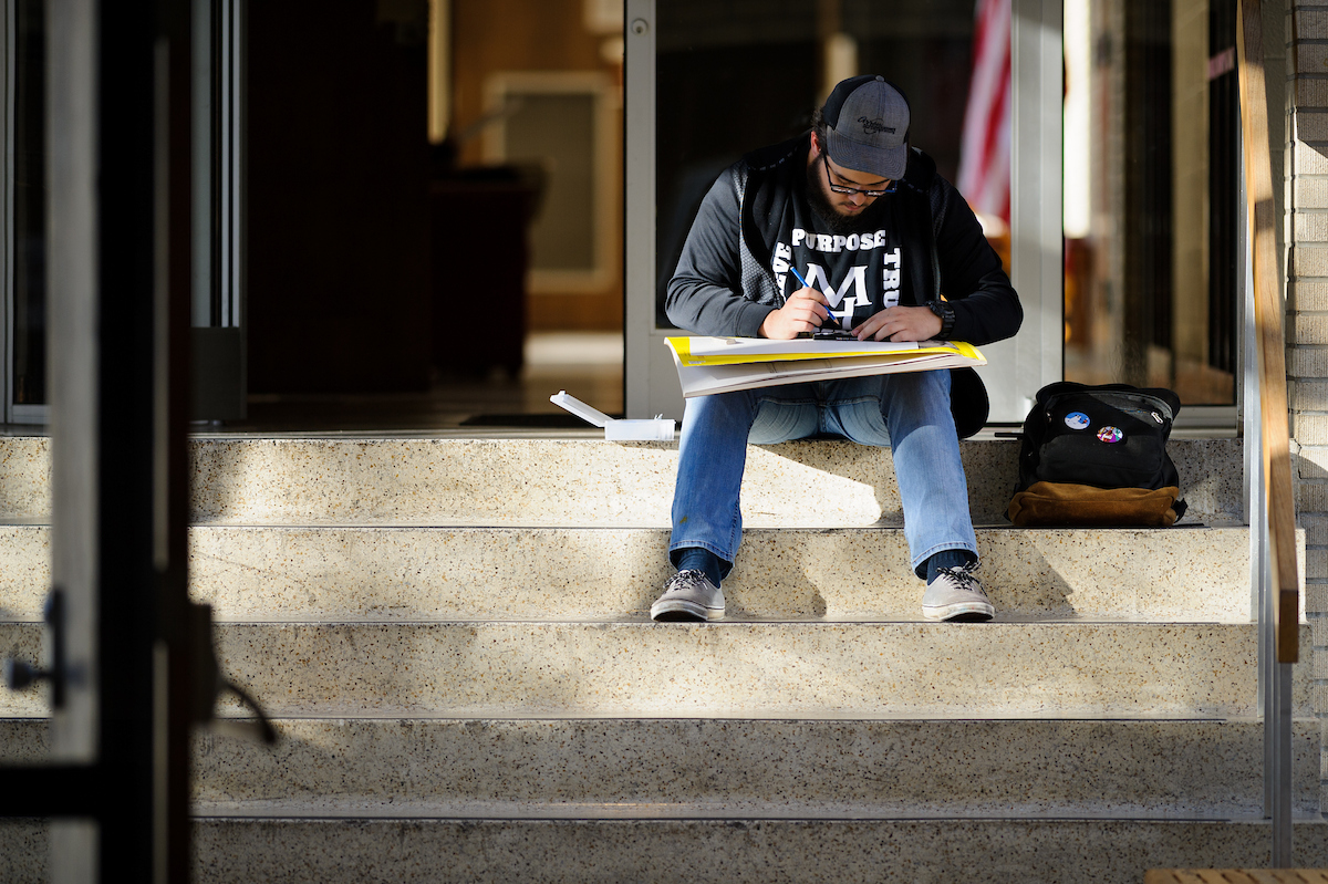 A-State student sits on campus steps focused on sketching in a large notebook.