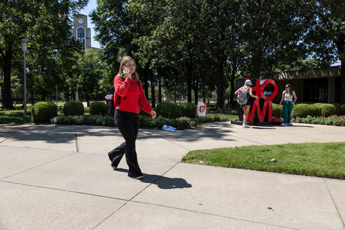 A student walks across campus while talking on the phone as two others pose by the red HOWL sign near the clock tower.