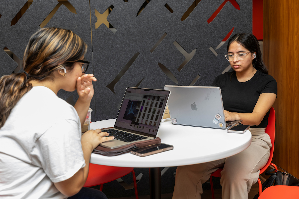 Two students studying with laptops at a round table indoors.