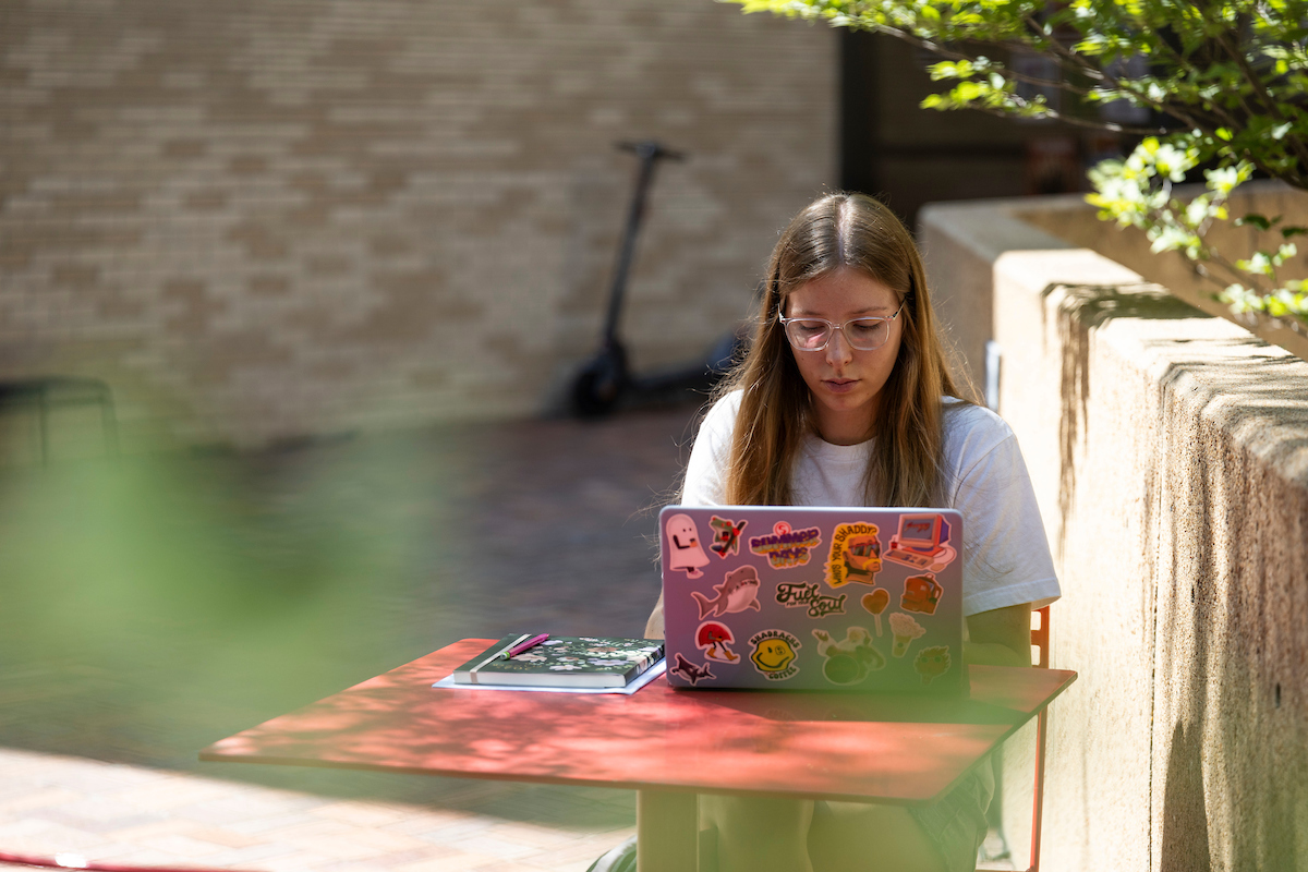 Student studies outdoors at ASU with a sticker-covered laptop and notebooks on a red table.