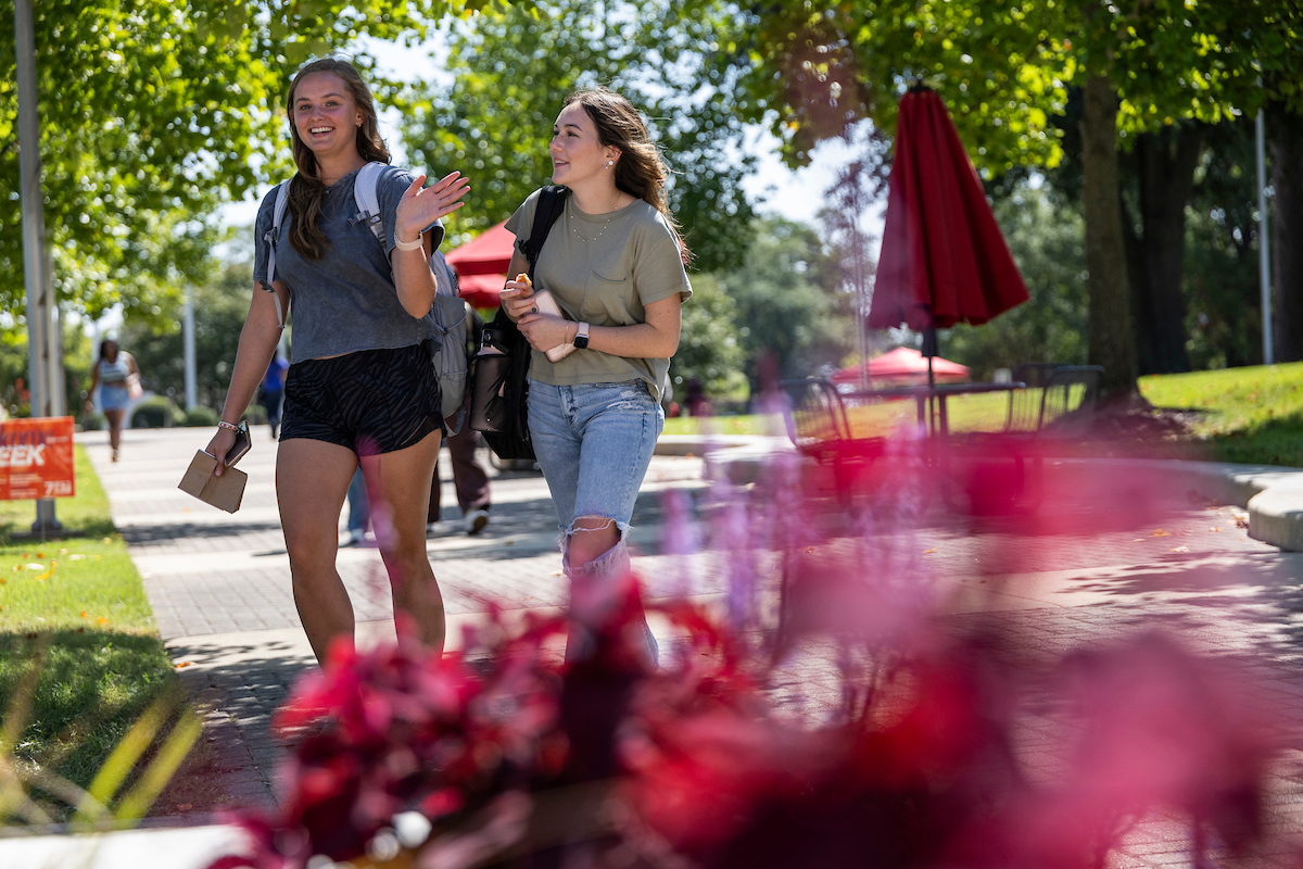 Two students walk and laugh on campus sidewalk with red umbrellas and summer flowers in the foreground.