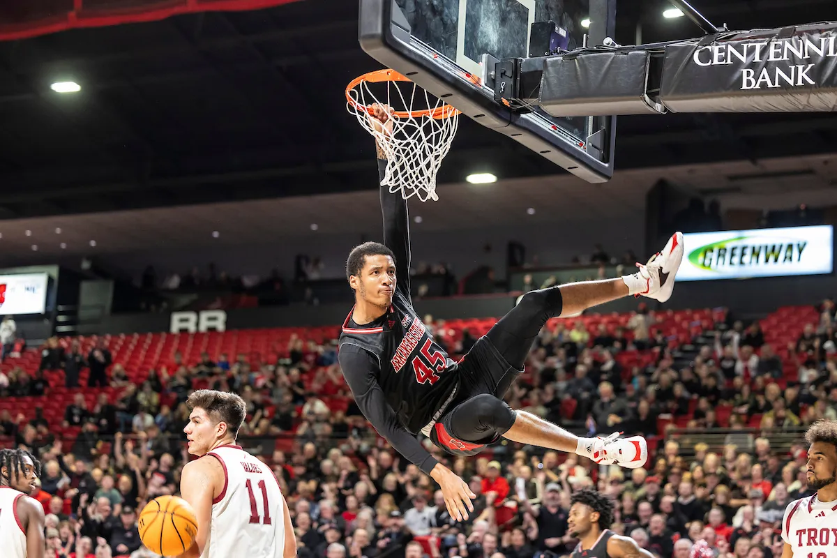 An A-State basketball player hangs on the rim after dunking over a Troy defender.