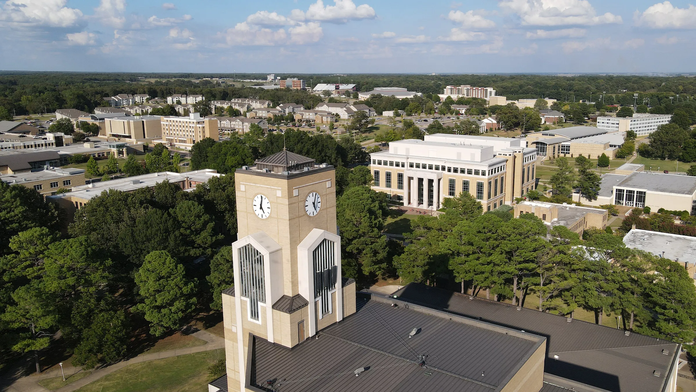 Aerial view of Arkansas State University campus featuring the iconic clock tower, surrounded by academic buildings, and tree-lined pathways under a cloudy blue sky