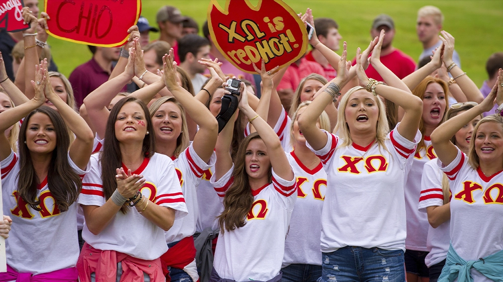 Group of people wearing Chi Omega shirts clapping and holding signs outdoors during Bid Day.
