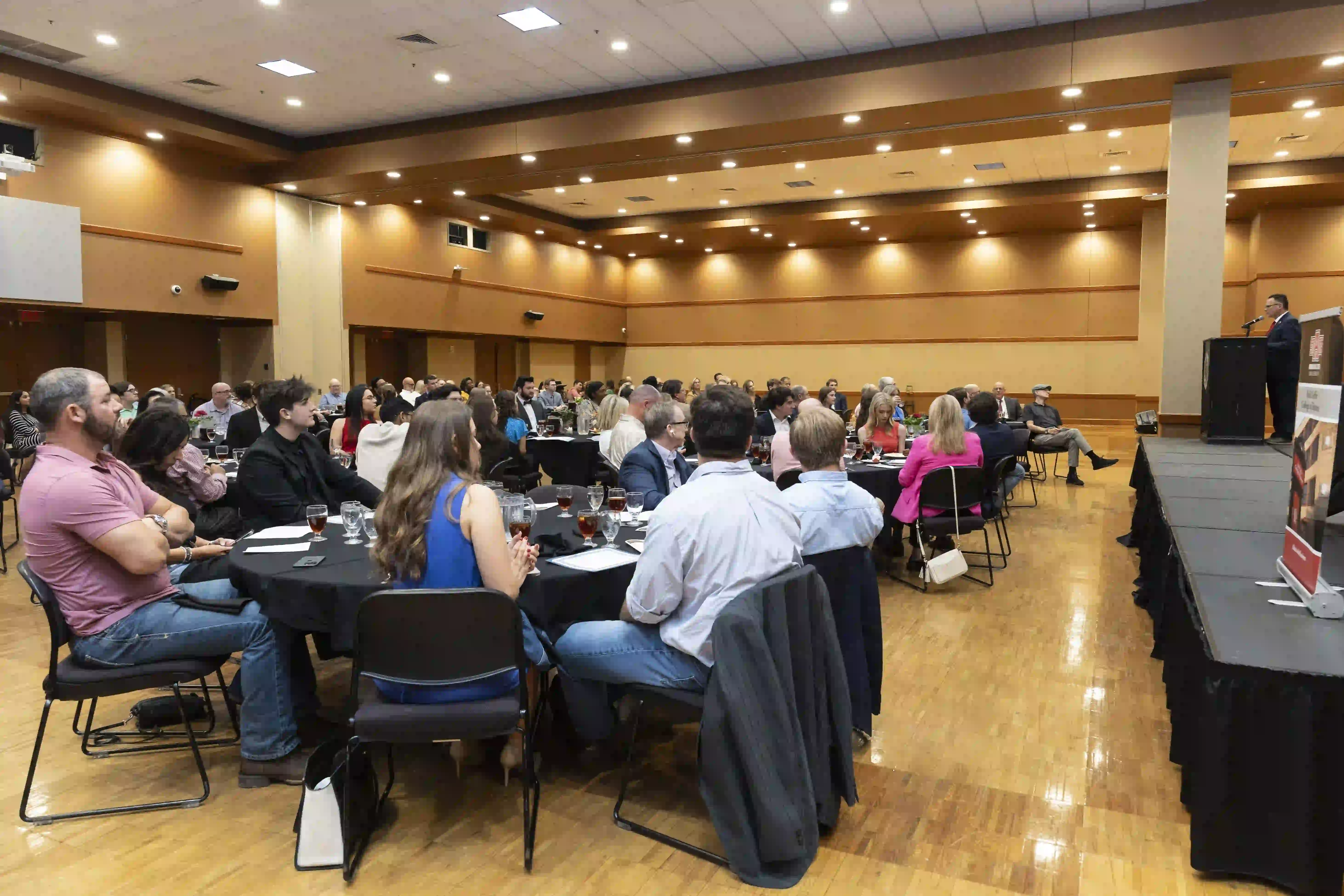 Students sitting at round tables facing a speaker on stage in Centennial Hall.