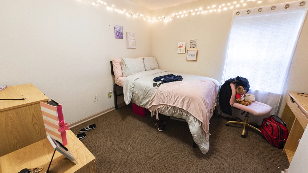 A bedroom with a pink comforter, string lights, and a wooden dresser. A chair and personal items add a lived-in feel.