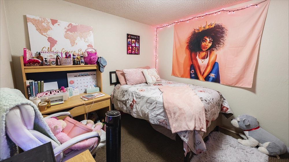 A bedroom decorated in pink tones with a wall tapestry, string lights, and a desk with books and supplies. A plush toy sits near the bed.