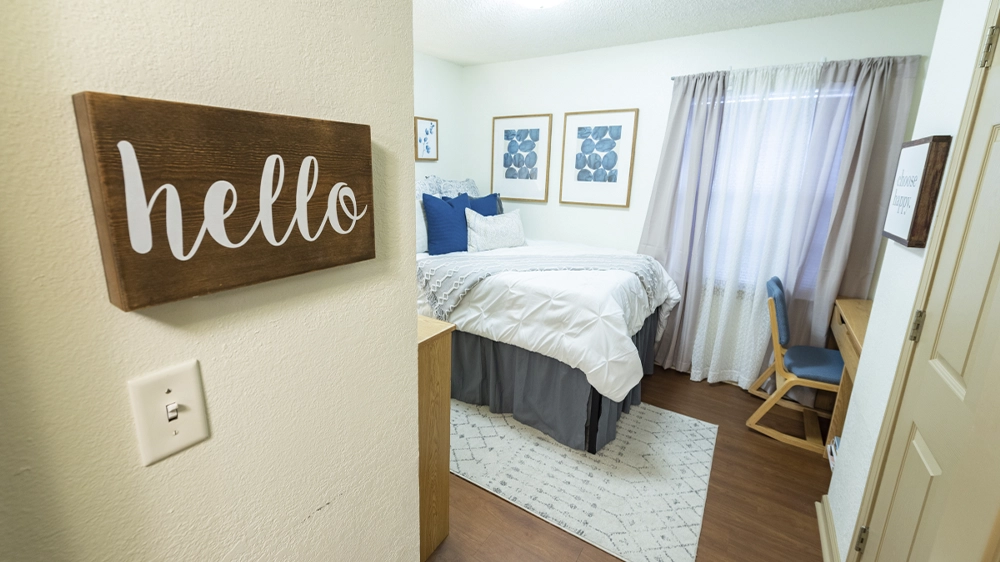 A bedroom with a white comforter, blue accent pillows, and framed artwork on the walls. A wooden desk and chair sit near the window with light curtains.